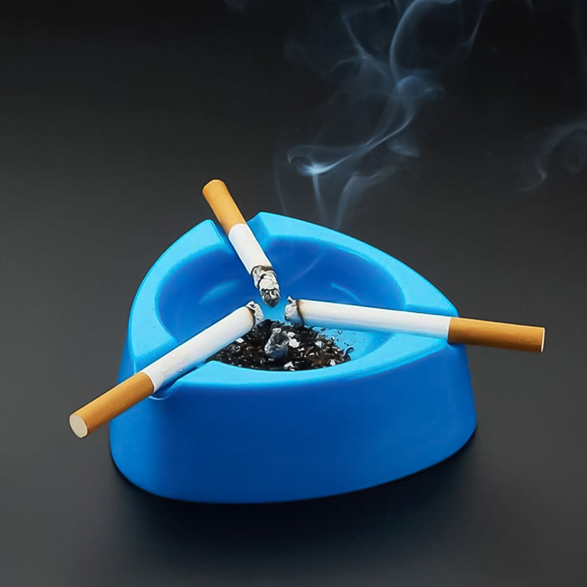 Three lit cigarettes rest in a blue plastic ashtray with ashes, emitting smoke on a dark surface.