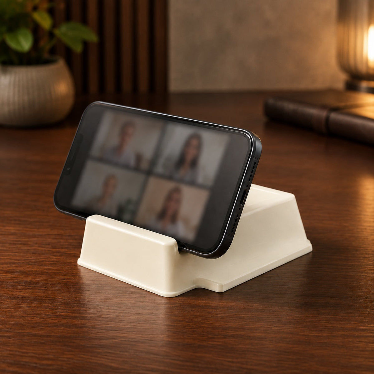 A black smartphone displaying a video call is propped horizontally in a cream plastic stand on a wooden desk.