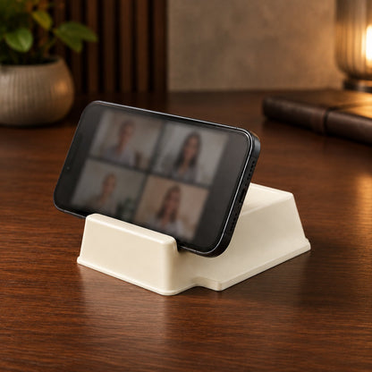 A black smartphone displaying a video call is propped horizontally in a cream plastic stand on a wooden desk.