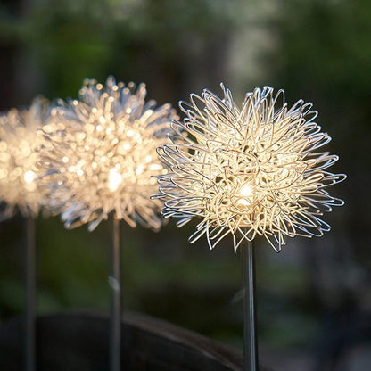 Three dandelion-shaped garden lights with warm white LEDs glow brightly in a soft-focus garden setting.