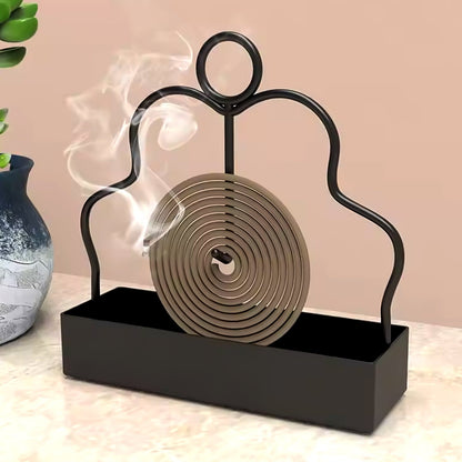 A black metal mosquito coil holder with a burning brown spiral coil emitting smoke next to a blue vase.
