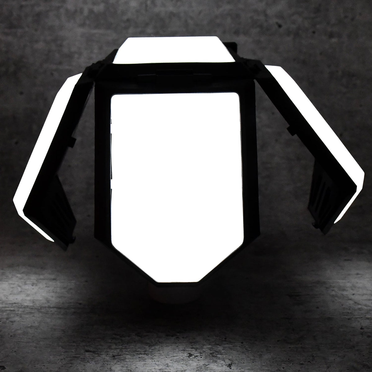 The white and black foldable LED lantern with its panels partially open, glowing brightly in a dark setting.