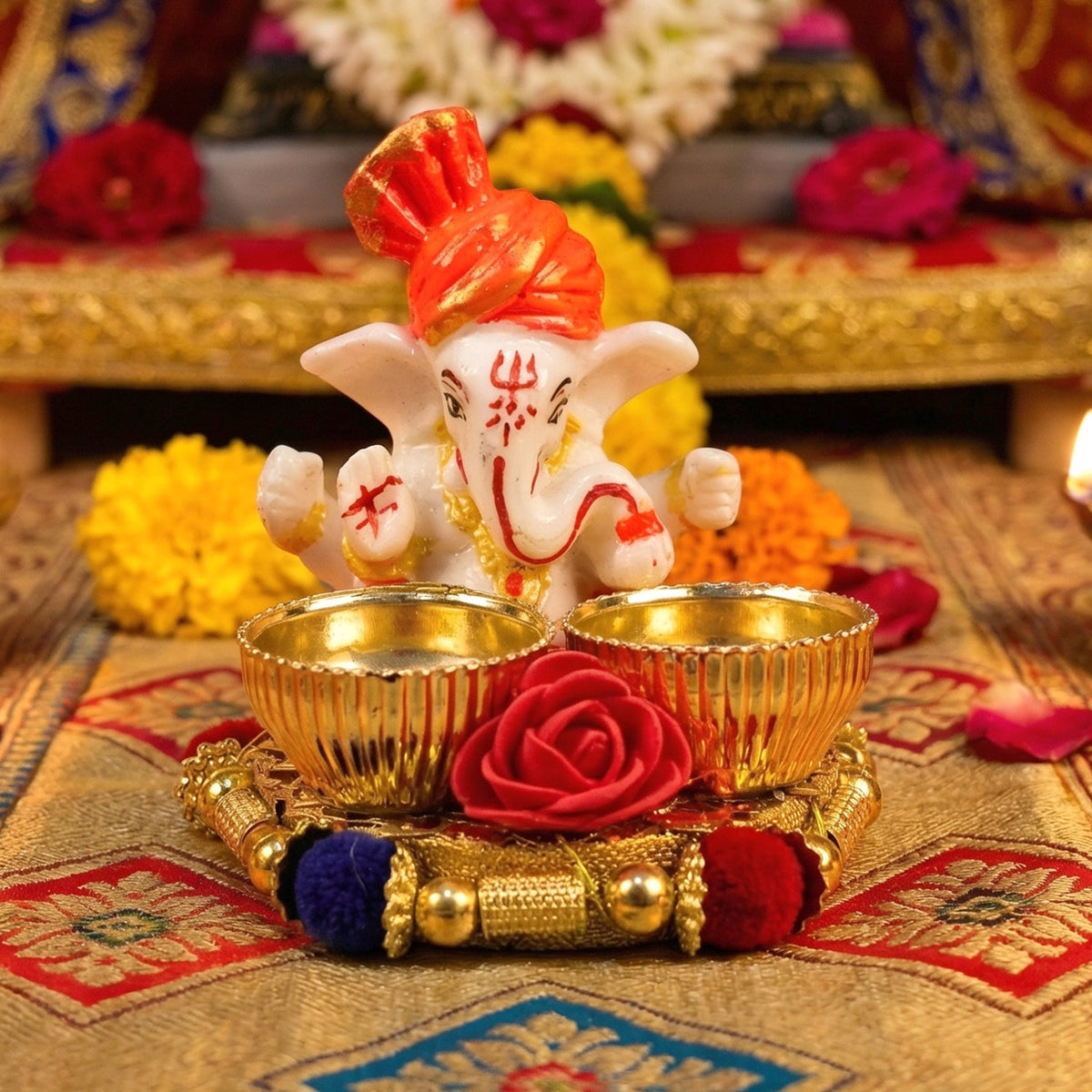 A white Ganesha idol with an orange turban on a gold base with two empty gold bowls and a red rose, on a gold patterned