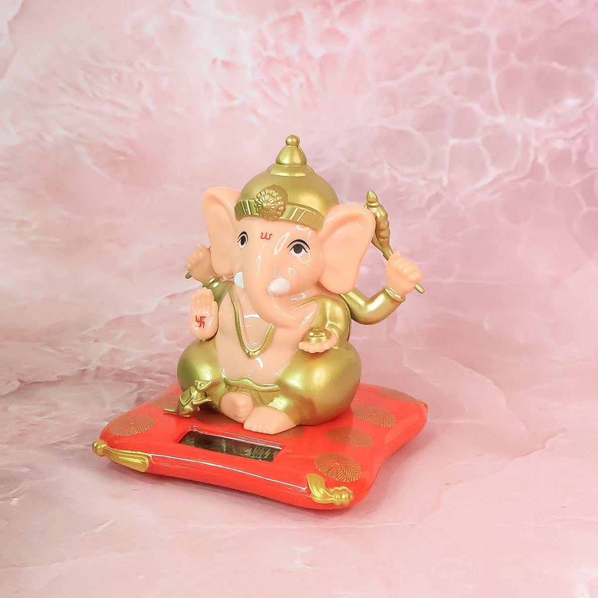 A Ganesha idol on a red base with gold accents, placed on a pink marble-patterned surface.