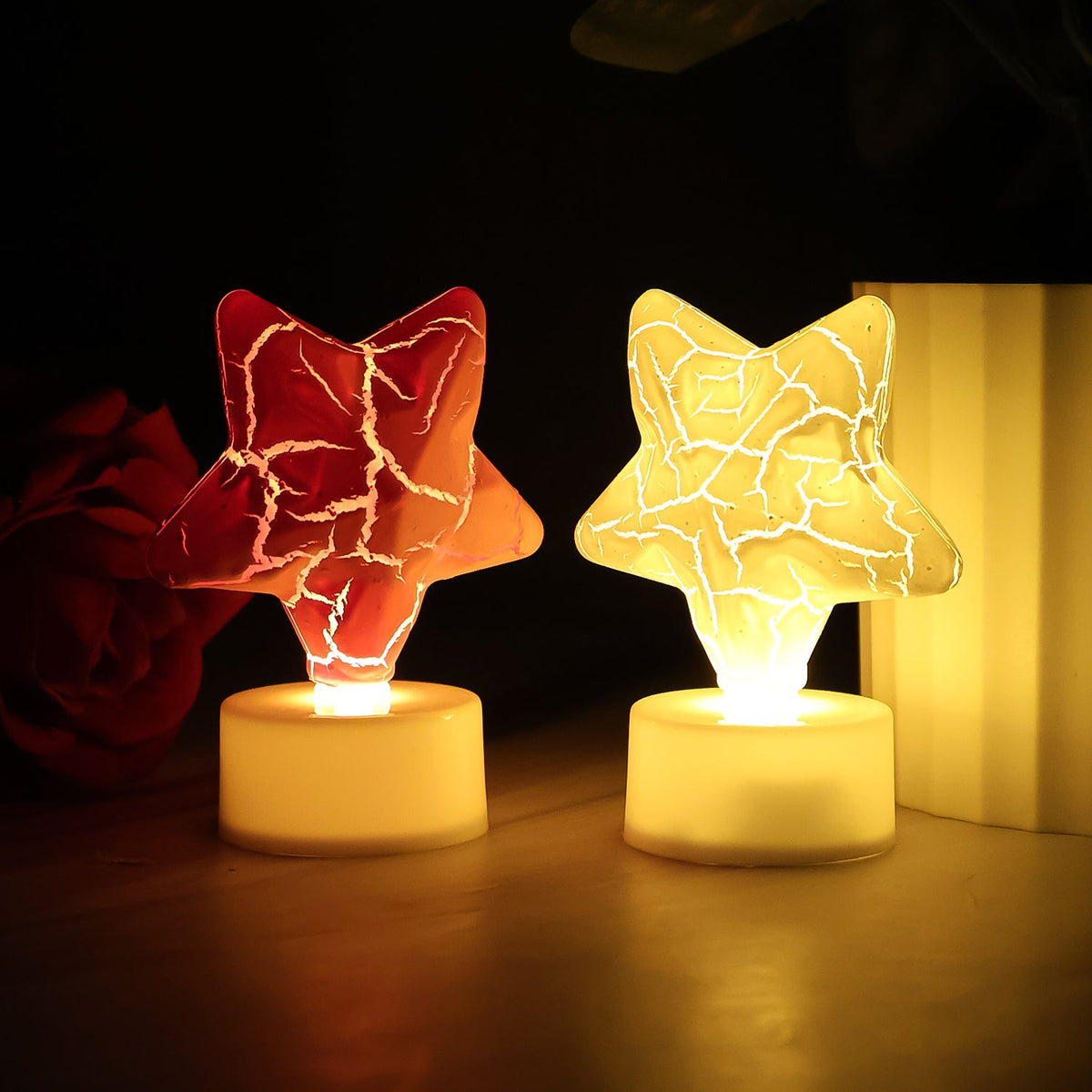 Two glowing star-shaped LED tealight candles, one red and one yellow, on a dark surface with a red rose in the background.