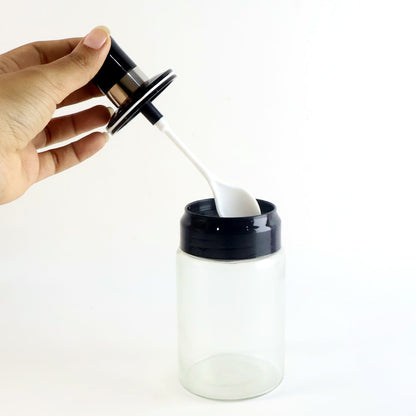 A hand holding the black lid and white spoon above a transparent spice jar.
