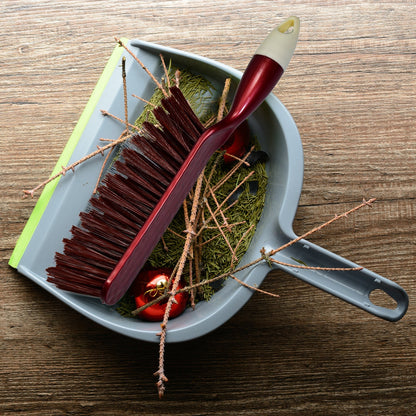 A red and white handled brush with brown bristles rests inside a gray dustpan filled with pine needles and small red
