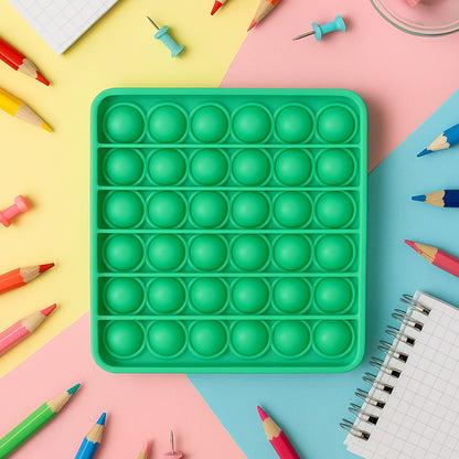 A square green silicone pop-it fidget toy surrounded by colorful pencils and thumbtacks on a multi-colored background.