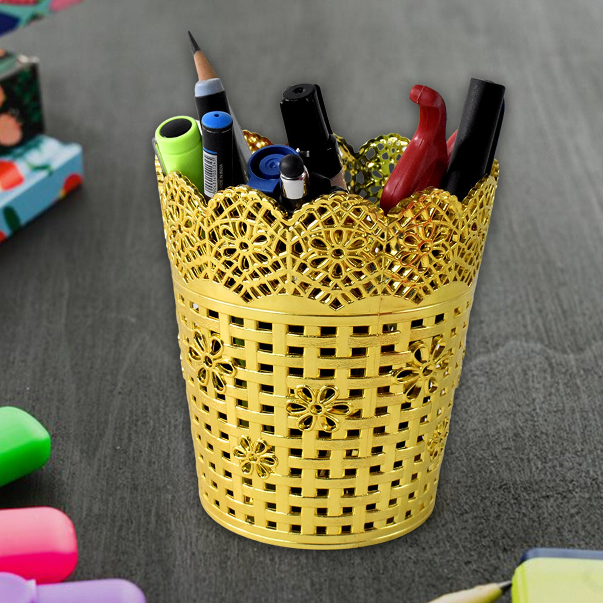 A gold plastic pen holder with a floral lace top rim is filled with various pens and markers on a dark textured surface.