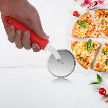 A hand uses a red-handled pizza cutter to slice a pizza with basil leaves on a white surface.