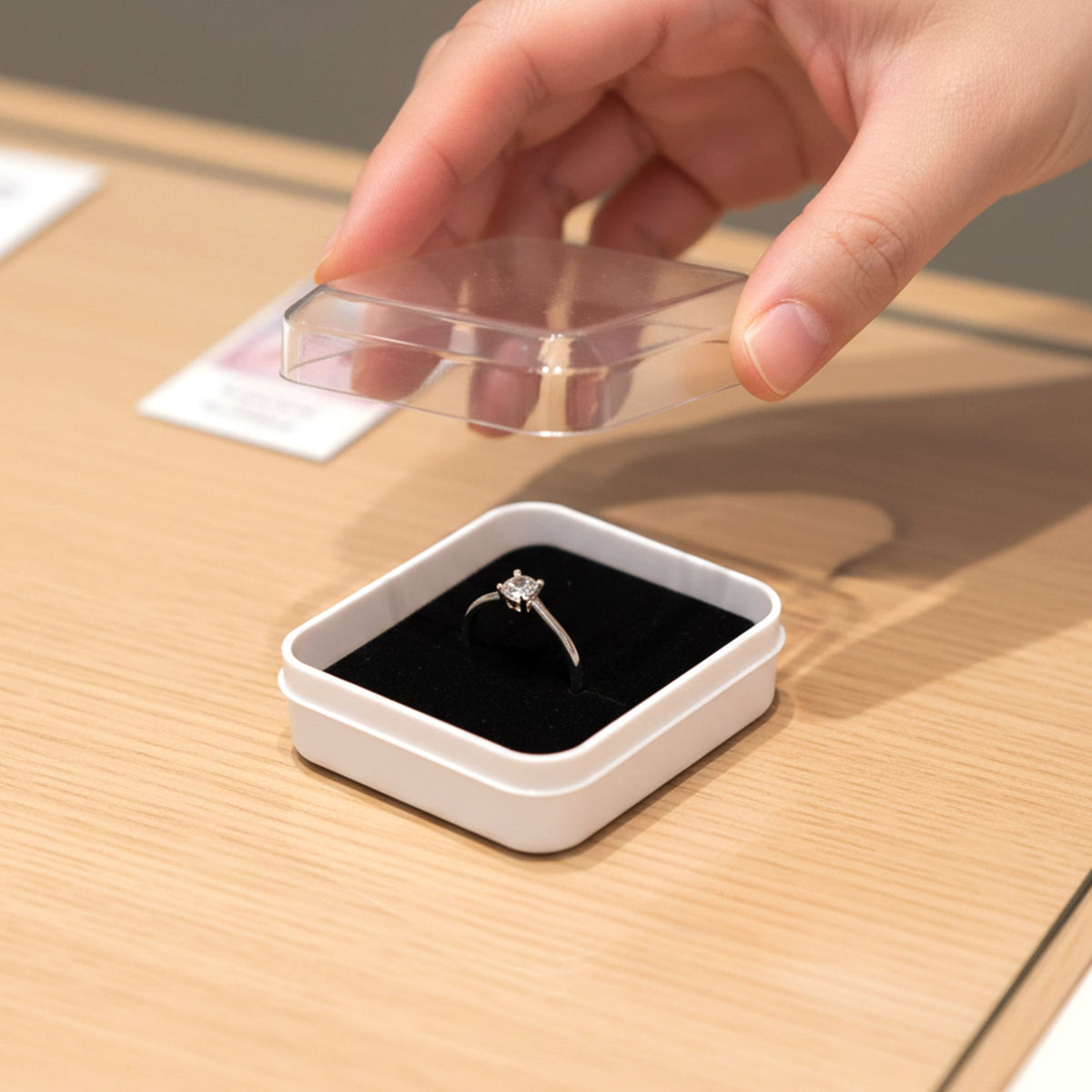 A hand opening a clear-lidded white jewellery box containing a silver ring with a square diamond on a light wooden table.