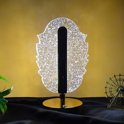 A glowing feather-shaped crystal table lamp with a gold base on a dark surface, with a hint of green foliage.