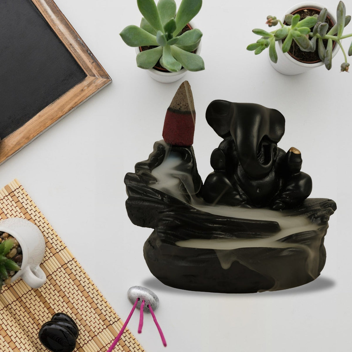 Overhead view of a black Ganesha smoke fountain with flowing smoke, surrounded by small potted succulents, a chalkboard, and
