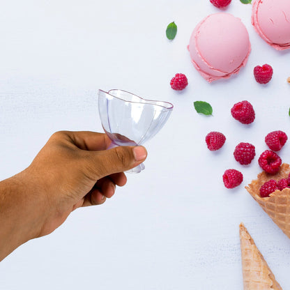 A hand holding a clear plastic ice cream bowl, with raspberries and pink ice cream in the background.