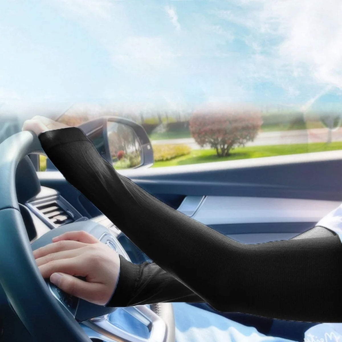 A person driving a car, wearing black compression sleeves on both arms, with a sunny outdoor view.