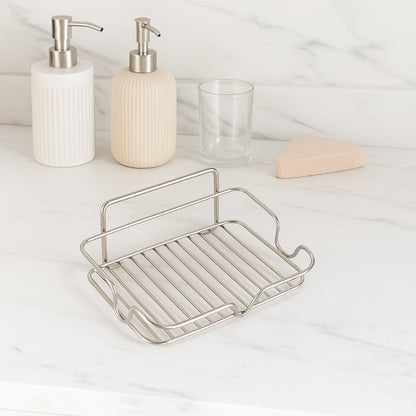 A stainless steel wire soap dish sits on a white marble counter with two soap dispensers and a glass in the background.