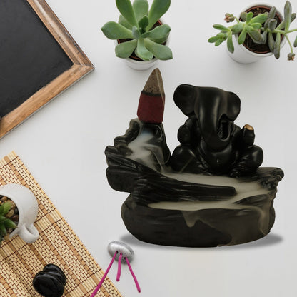 A black Ganesha backflow incense burner with smoke, succulents, and a small mat on a white surface.