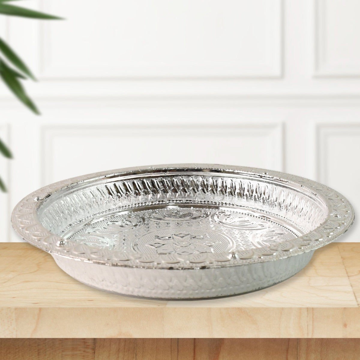 A silver-colored plastic pooja thali with embossed patterns, sitting on a light wooden surface with a white paneled wall
