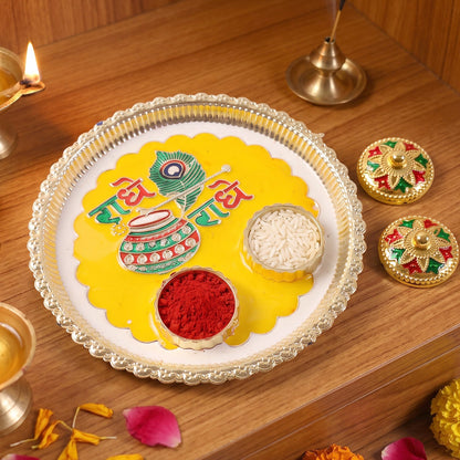 A silver and yellow Radhe Radhe pooja thali with small containers of kumkum and rice, next to two decorative containers.