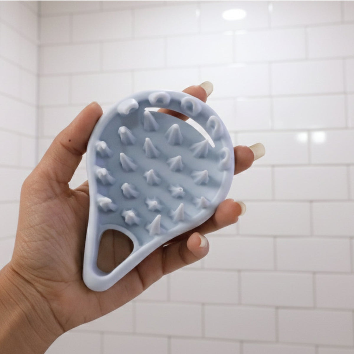 A hand holding the light blue silicone scalp massager, showing its spiky underside against a white tiled wall.