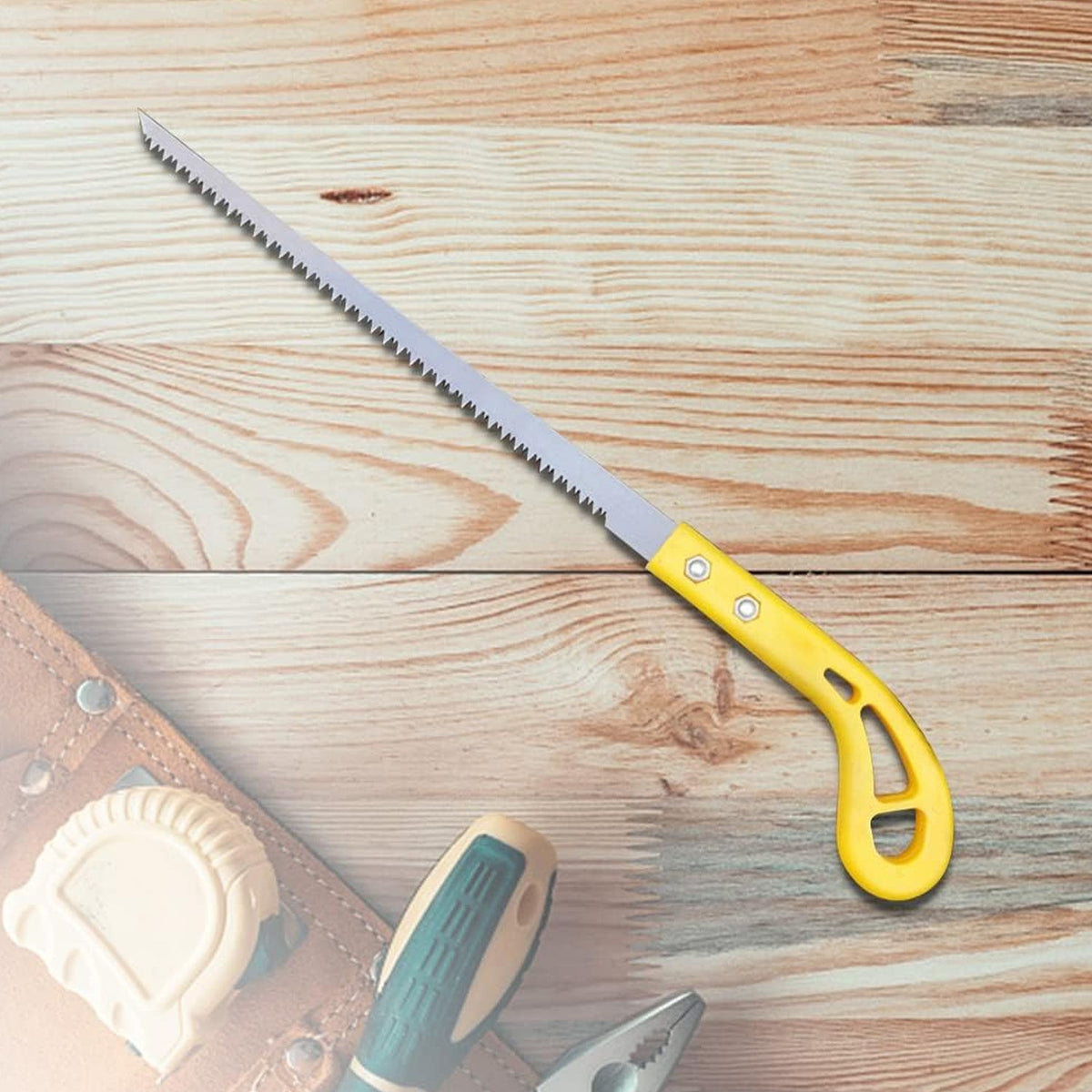 A yellow-handled pruning saw is displayed on a light wooden surface with blurred tools in the foreground.