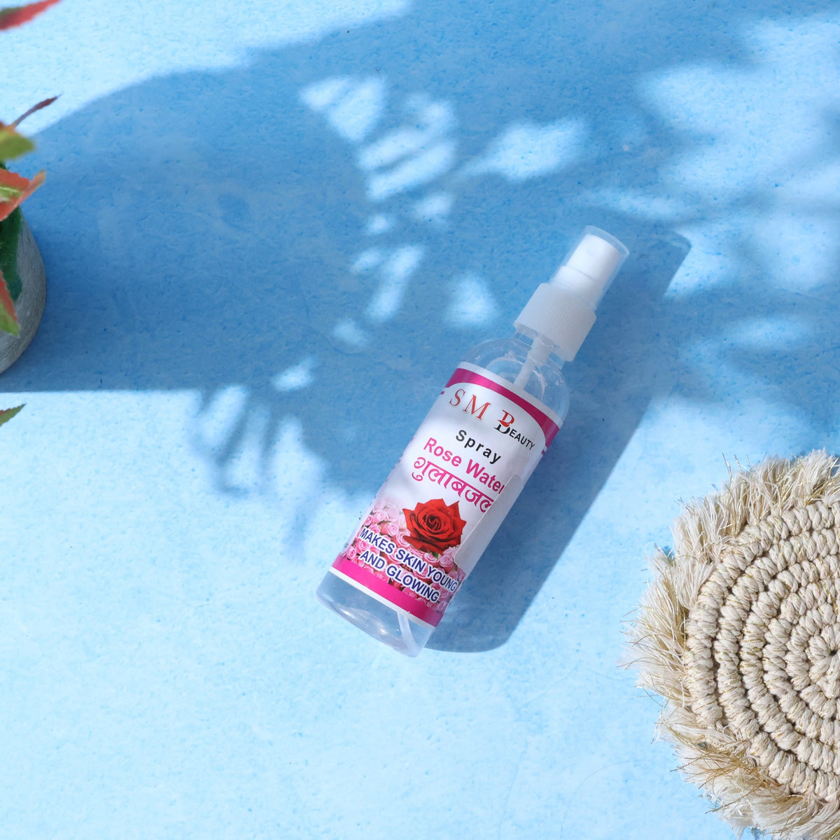 A clear bottle of SM Beauty rose water spray with a pink label lies diagonally on a blue surface with shadows.