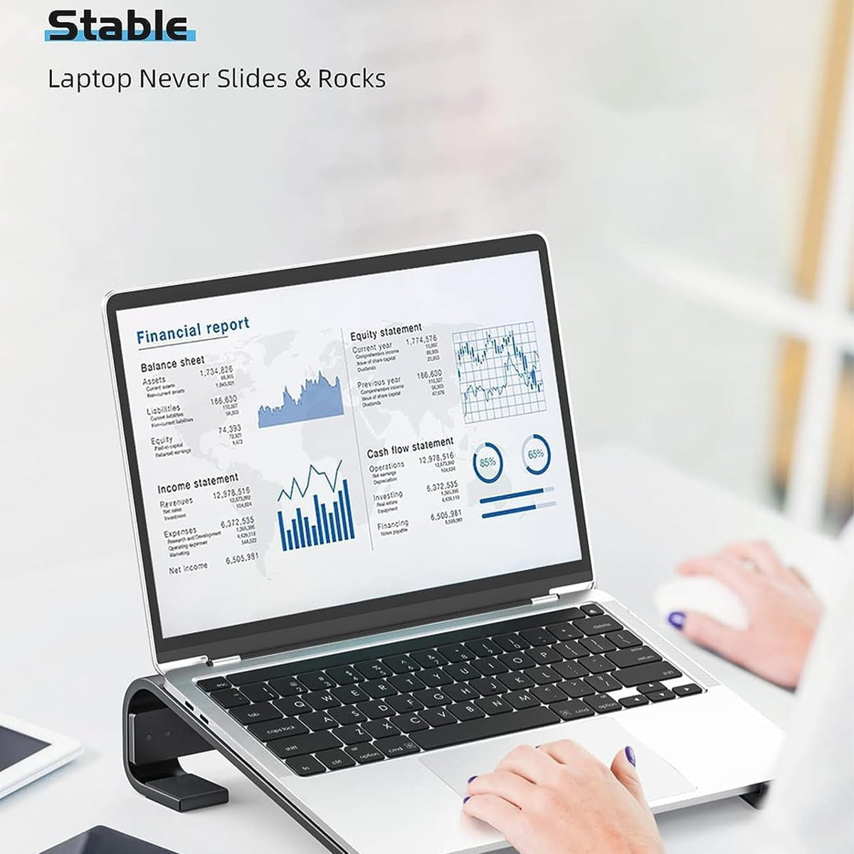 A person's hands type on a silver laptop resting on a black stand, displaying a financial report.