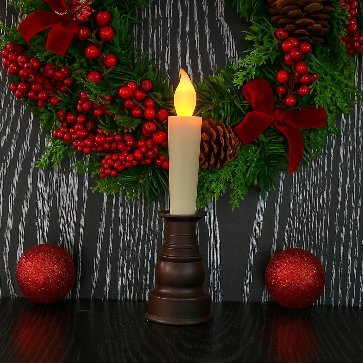 A cream LED candle in a dark bronze holder in front of a festive wreath with red berries and bows.