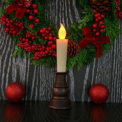 A cream LED candle in a dark bronze holder in front of a festive wreath with red berries and bows.