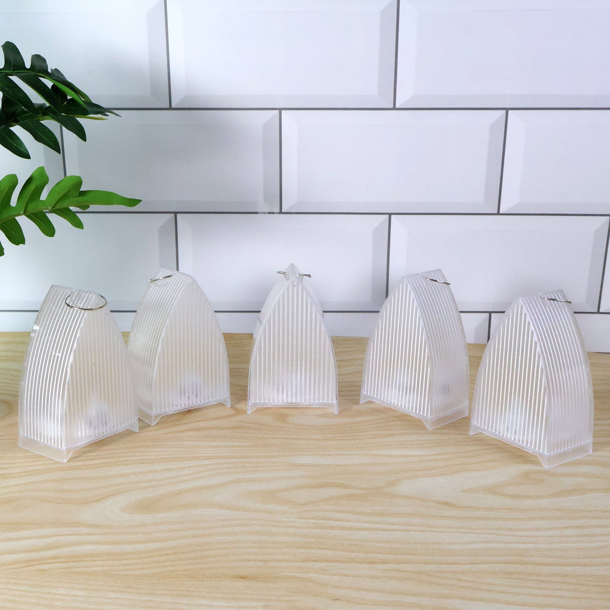 Five clear plastic ribbed lanterns with metal hooks on top, arranged on a light wooden surface against white subway tiles.