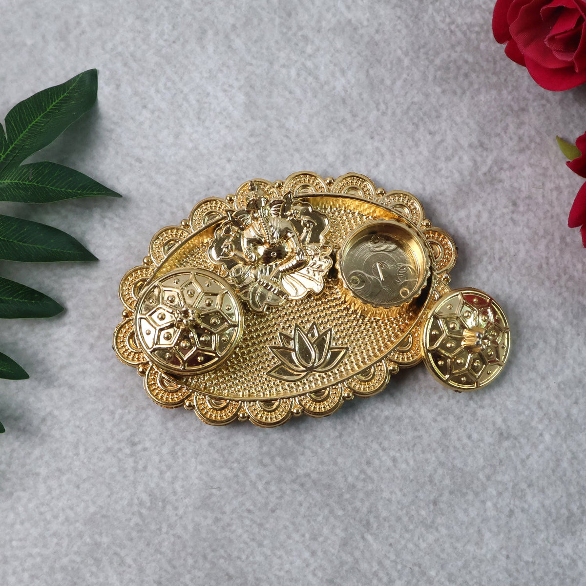 A gold-finished chopra with Ganesh, two containers, and a lotus design on a grey textured surface with green leaves and red