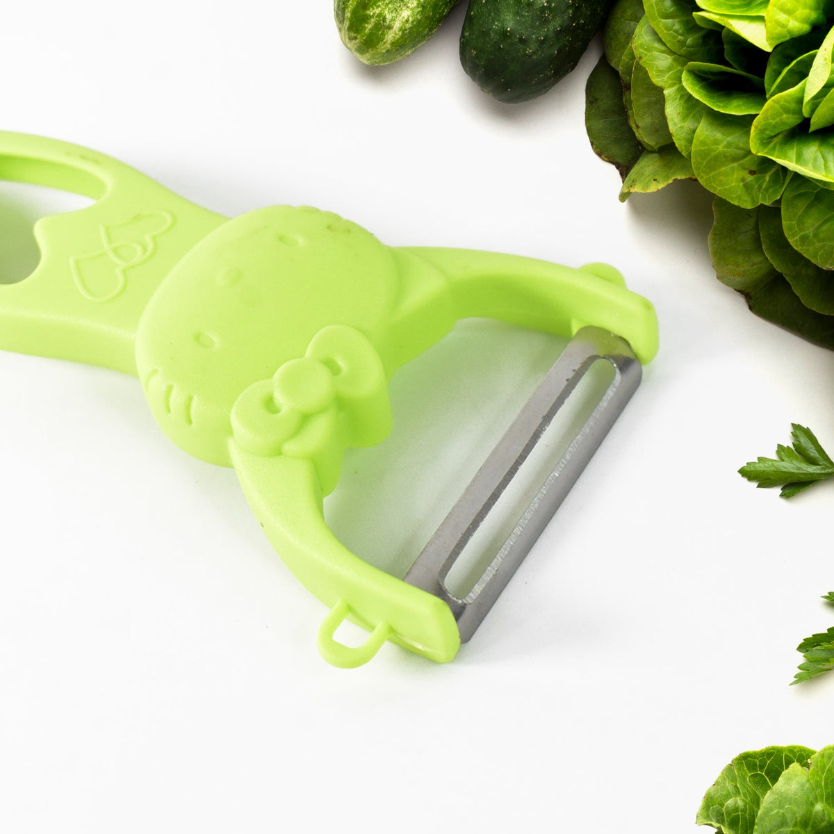 Close-up of a green peeler with a Hello Kitty design and a stainless steel blade, next to fresh greens.