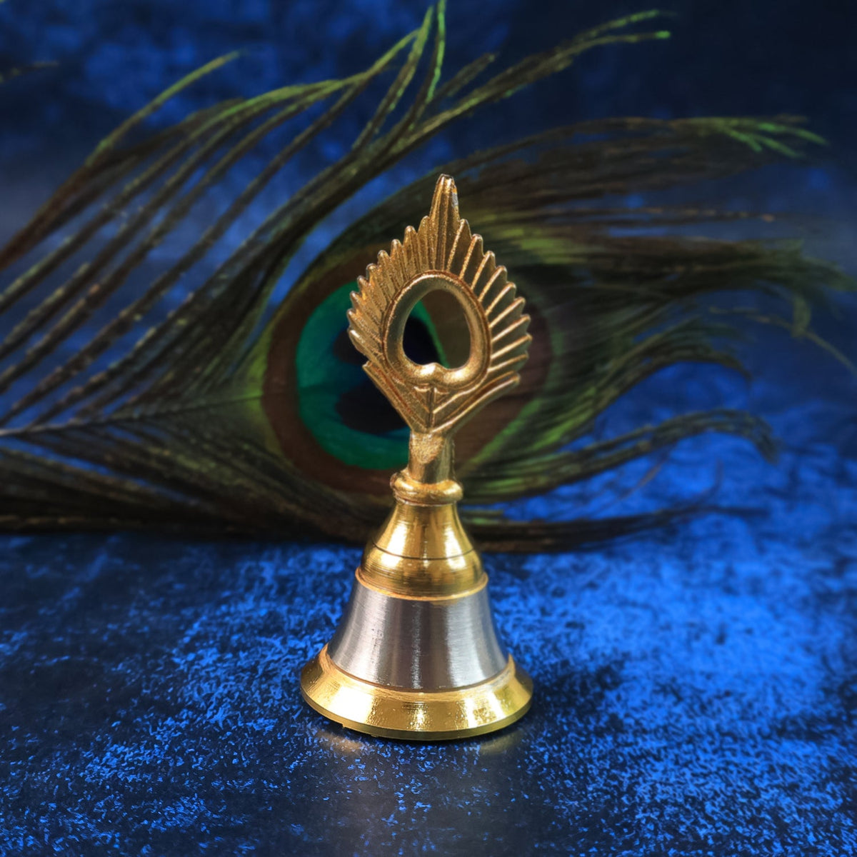 A small brass and steel pooja bell with a peacock feather design sits against a dark blue background with a peacock feather.
