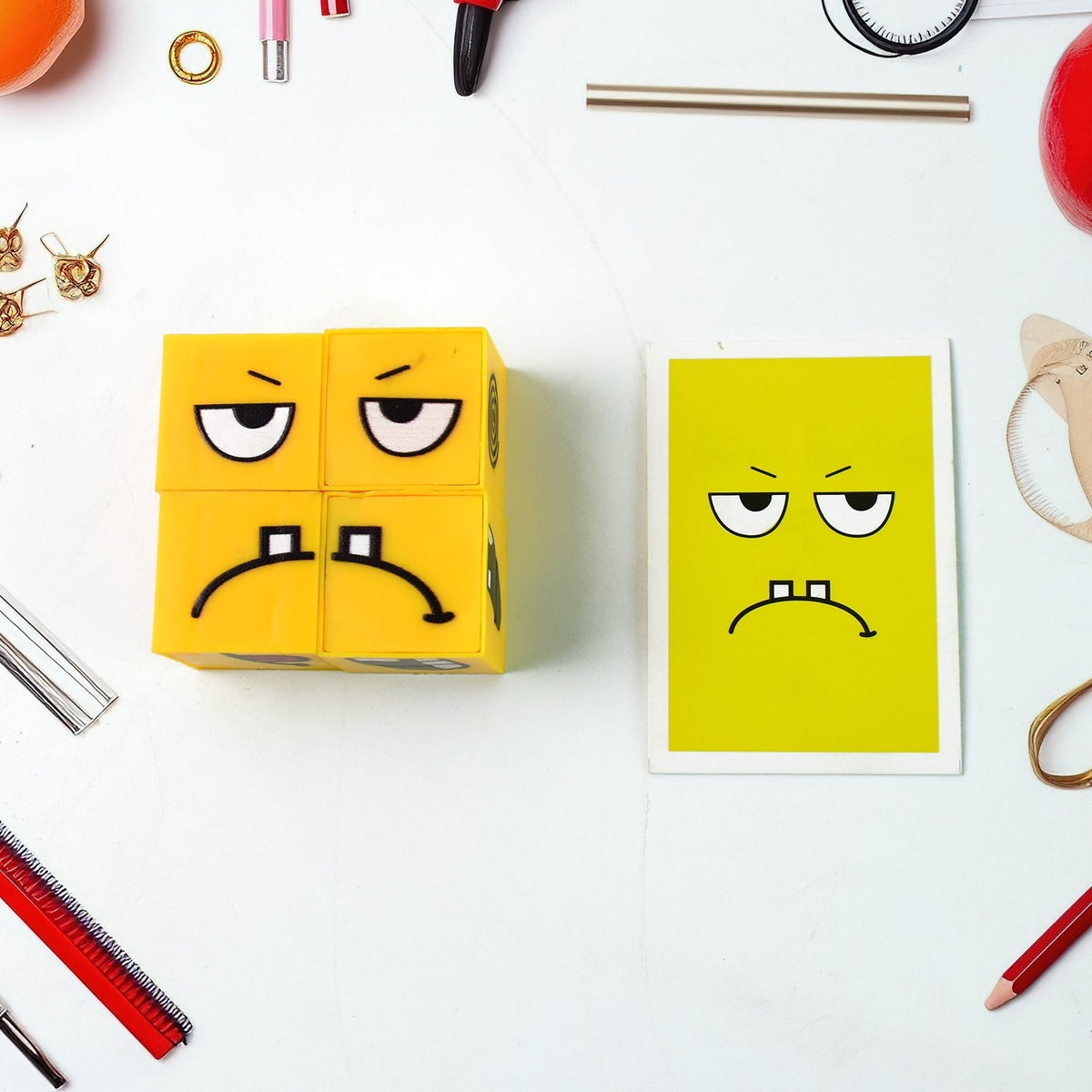 A yellow emotion cube displaying a sad face is next to a matching yellow card on a white surface with various office