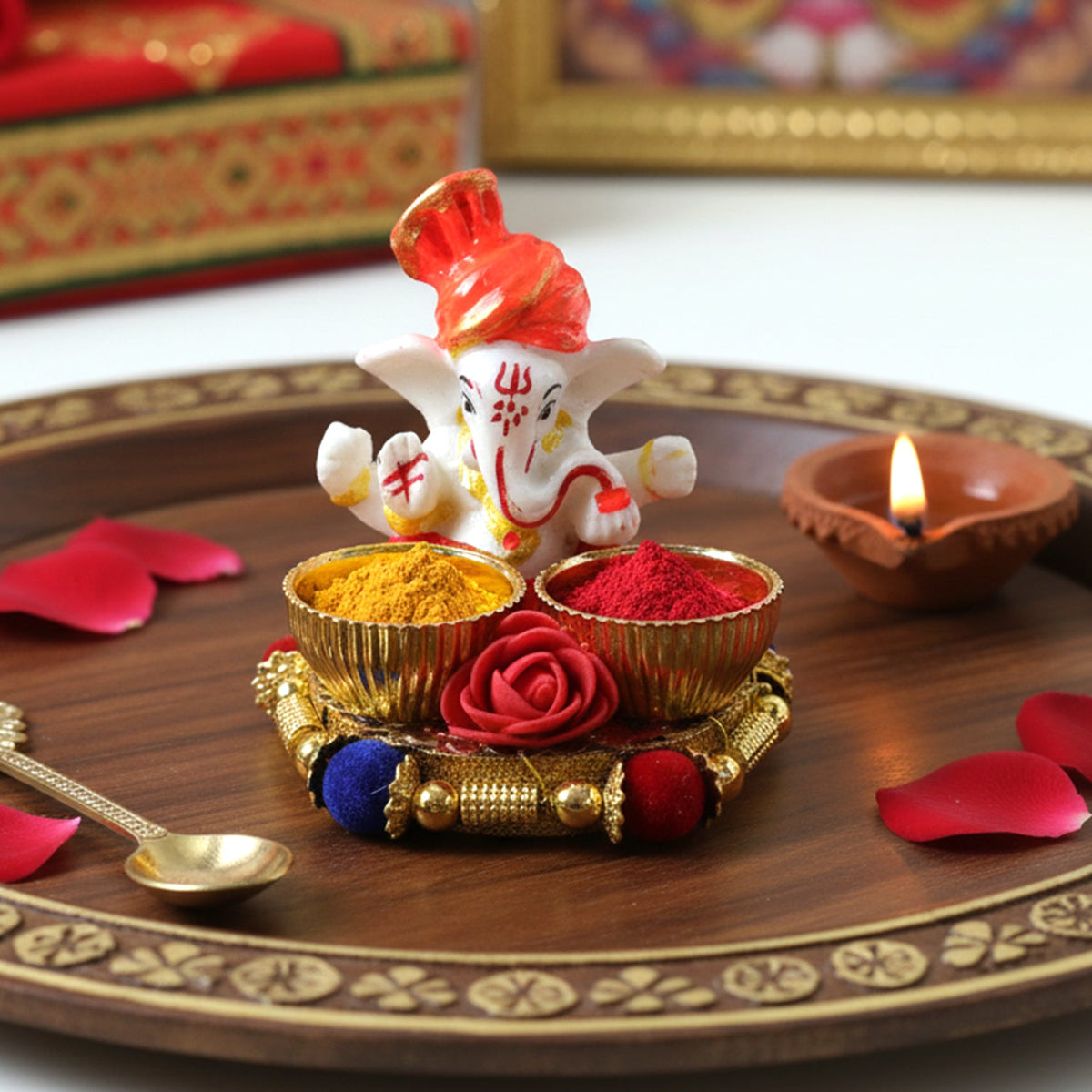 A white Ganesha idol with an orange turban on a gold base with two bowls filled with yellow and red powder, next to a lit