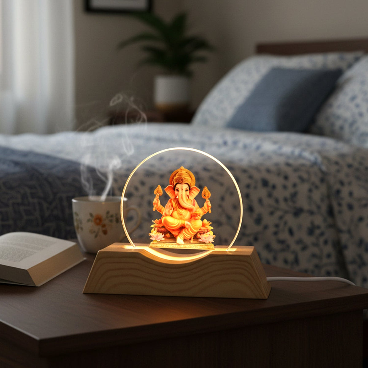Lord Ganesha LED lamp with a golden glow on a bedside table with a book and steaming mug.