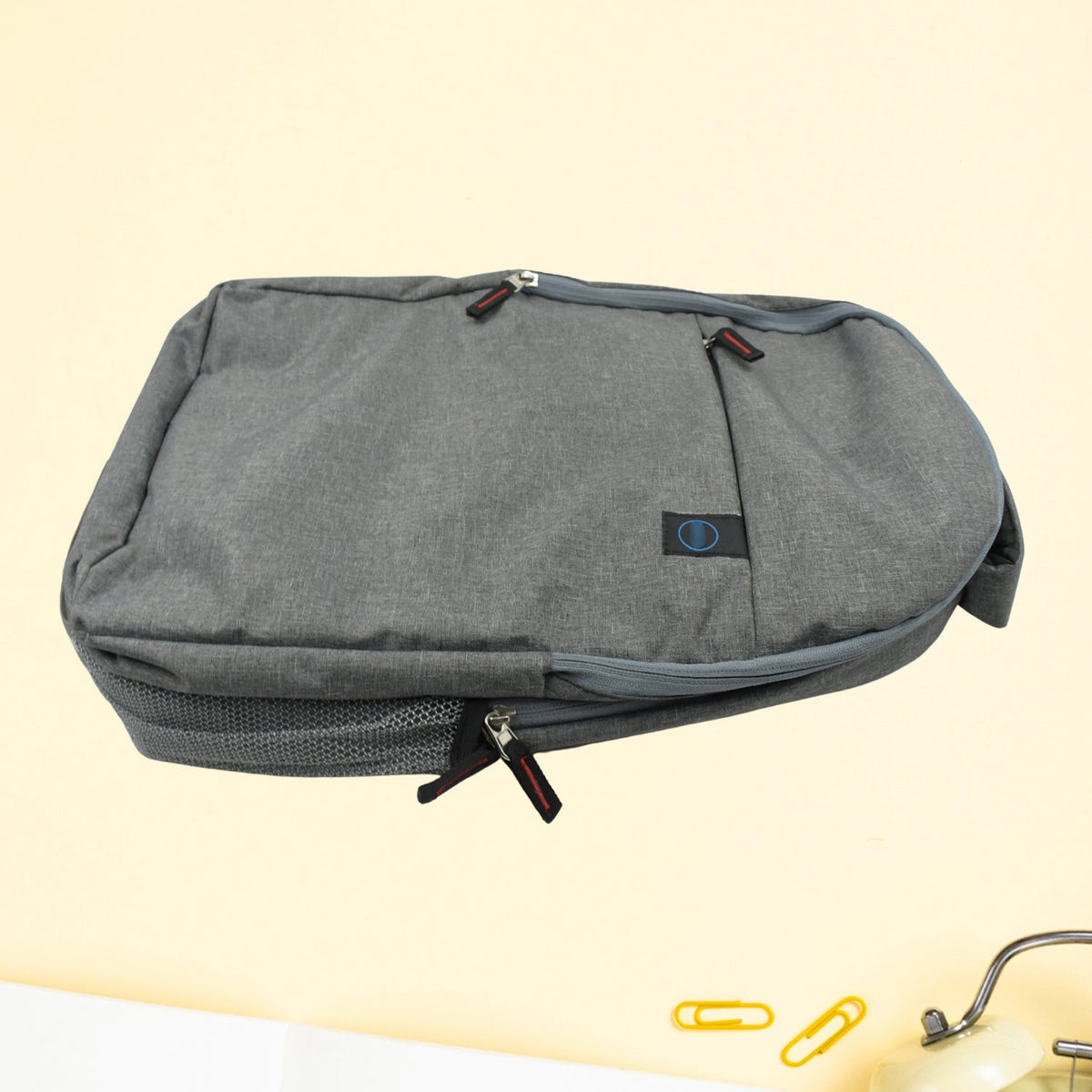 A grey backpack with multiple zippers and a mesh side pocket is shown on a light yellow background.