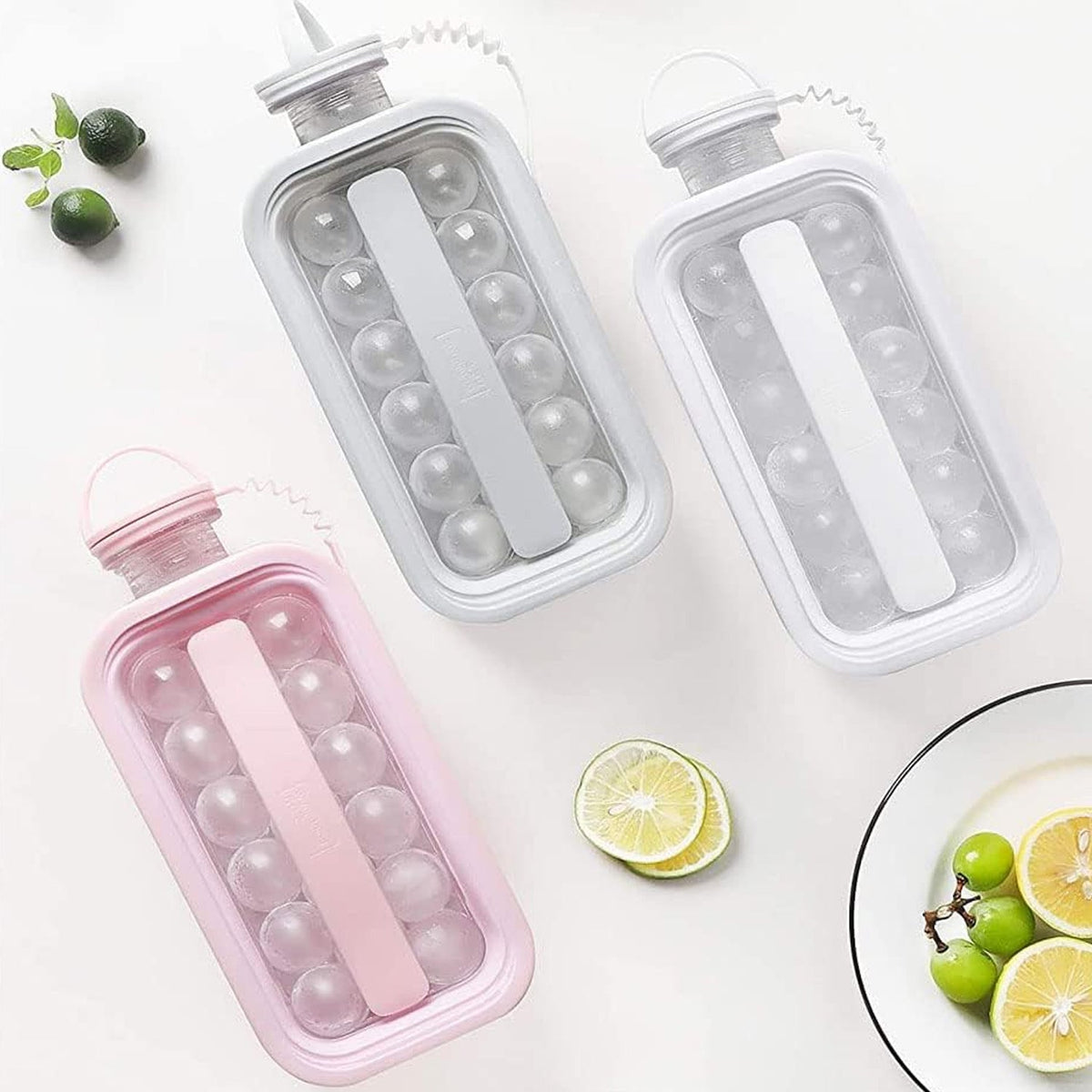 Three ice ball maker bottles in pink, grey, and white are arranged on a white surface with sliced citrus fruits and grapes.