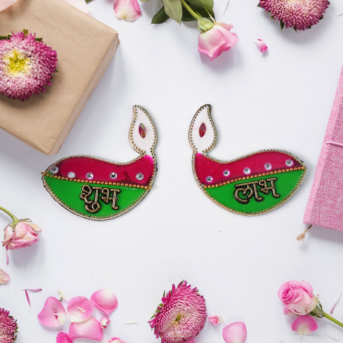 Two decorative diya stickers, pink and green with gold text, surrounded by pink flowers, petals, and a brown gift box.