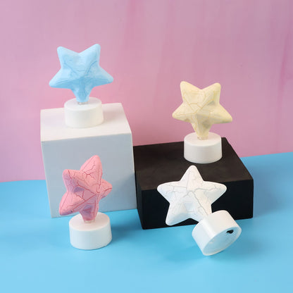 Four star-shaped LED tealight candles in blue, yellow, pink, and white, unlit, on blocks against a pink and blue background.