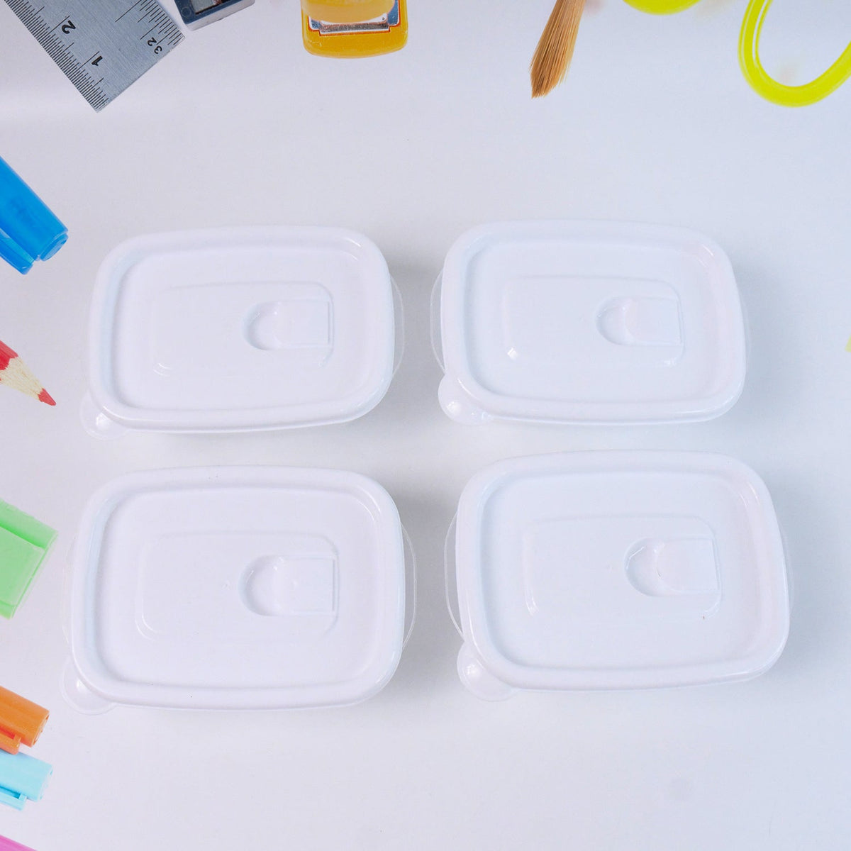 Top-down view of four clear plastic food containers with white lids on a white surface, surrounded by art supplies.