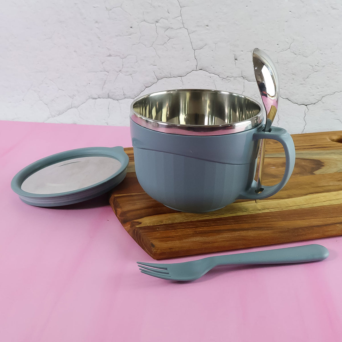 A blue insulated bowl with a lid, a spoon, and a fork on a wooden board with a pink background.