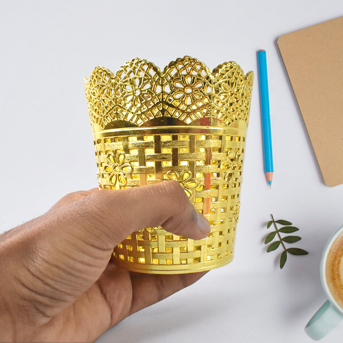 A hand holds a gold plastic pen holder with a floral lace top rim over a white desk with a pencil and notebook.