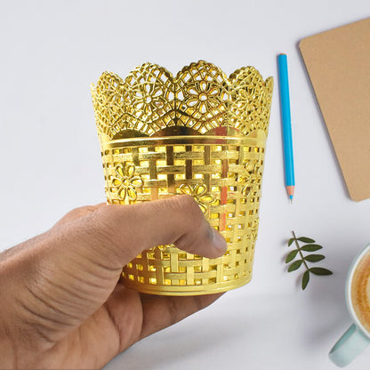 A hand holds a gold plastic pen holder with a floral lace top rim over a white desk with a pencil and notebook.