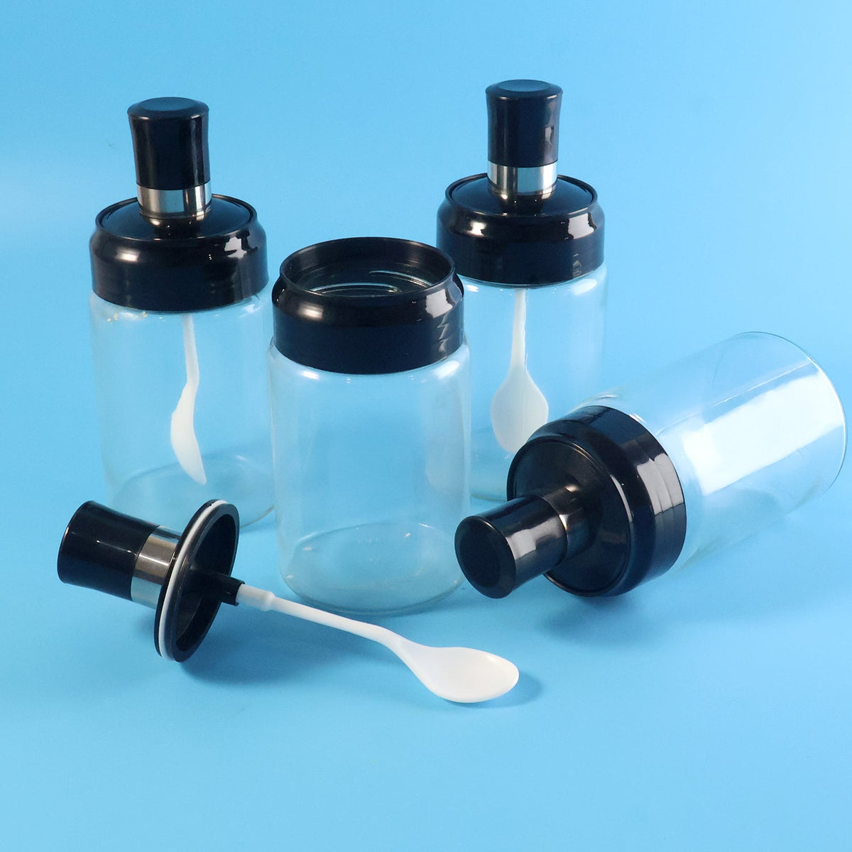 Four transparent spice jars, one with its black lid and white spoon detached on a blue background.