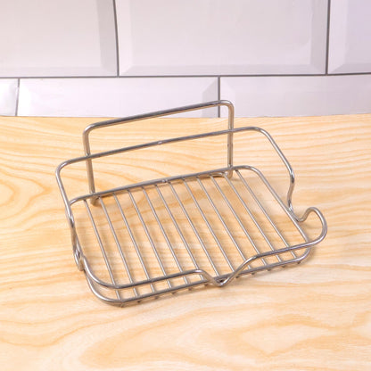 A stainless steel wire soap dish rests on a light wood surface with white subway tiles in the background.
