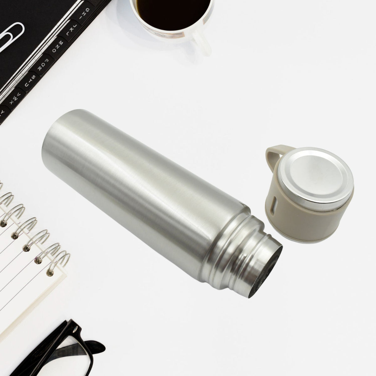 The silver vacuum flask lies on a white desk next to its detached beige cap, with a notebook and glasses nearby.