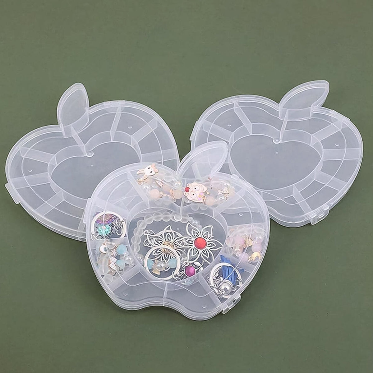 Three transparent apple-shaped organizer boxes are displayed on a dark green surface; the front box is open and filled with