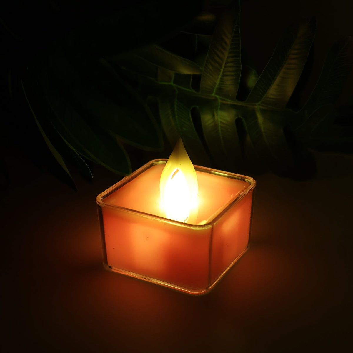 A single square LED flameless tealight candle glowing orange, with a dark background and green foliage.