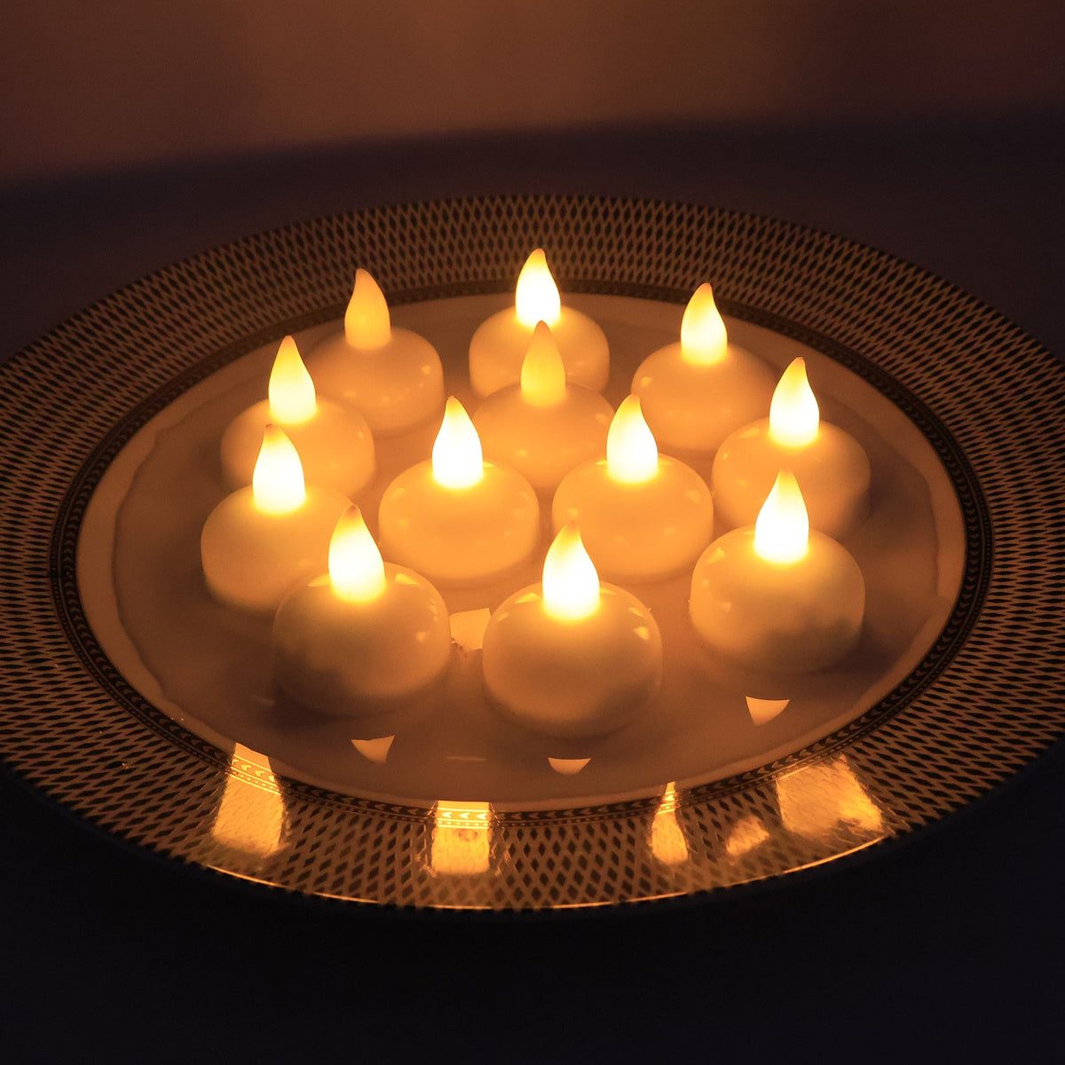Twelve white LED floating candles with warm yellow flames arranged on a decorative plate with a patterned rim.